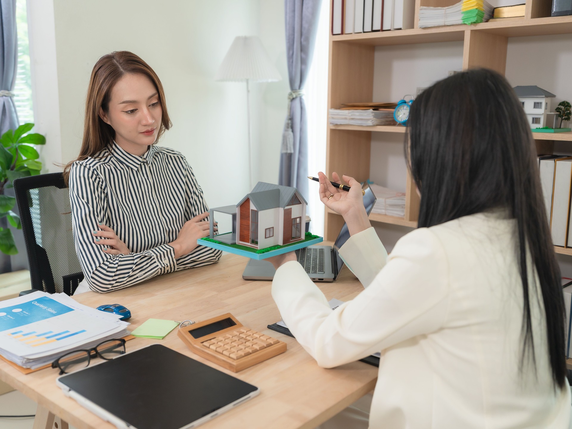 Two Asian female real estate businesspeople working together are discussing, focusing on a house plan on the table, suggesting a planning meeting or sales meeting.